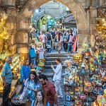 A view through a historic stone archway into a bustling alley of Khan El-Khalili bazaar in Cairo, filled with glittering traditional lanterns, brassware, and colorful artisanal crafts.