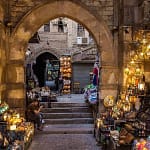 A view through a historic stone archway into a bustling alley of Khan El-Khalili bazaar in Cairo, filled with glittering traditional lanterns, brassware, and colorful artisanal crafts.