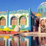 A view from the Nile River of a vibrant Nubian village in Aswan, showcasing traditional brightly painted blue and white houses along the rocky shoreline.