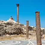 The ancient Roman Pompey's Pillar alongside a granite Sphinx statue under a blue sky in Alexandria Egypt.