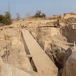 A high-angle view looking down at the massive Unfinished Obelisk carved directly into the pink granite bedrock of an ancient quarry in Aswan, under a clear sky. Caption A fascinating glimpse into ancient craftsmanship: the monumental Unfinished Obelisk