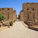 The stone pathway leading to the massive pylon entrance of Karnak Temple in Luxor, lined on both sides by ancient ram-headed sphinx statues under a bright blue sky.