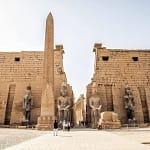 The grand entrance pylon of Luxor Temple with ancient stone obelisks and statues under a bright sky.