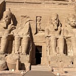 A front-facing daytime view of the Great Temple of Abu Simbel, showcasing the towering carved stone colossi statues of Pharaoh Ramesses II guarding the rock-cut entrance.