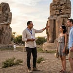 A professional private Egyptologist in smart-casual attire guiding a couple in front of the massive Colossi of Memnon in Luxor.