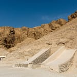 A wide daytime view of the desert landscape at the Valley of the Kings in Luxor, showing two concrete-shaded ramps leading down into underground ancient tombs carved into the limestone hills.