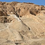 A wide daytime shot focusing on the rugged, sandy limestone cliffs of the Valley of the Kings in Luxor, showing a dirt path leading up to ancient rock-cut tomb openings on the hillside.