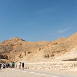 A wide daytime view of the arid, rocky desert hills of the Valley of the Kings in Luxor, with a group of tourists walking along a paved visitor pathway.