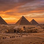 Wide-angle panoramic view of the three Great Pyramids of Giza and the Sphinx under a vibrant orange and purple sunset sky.