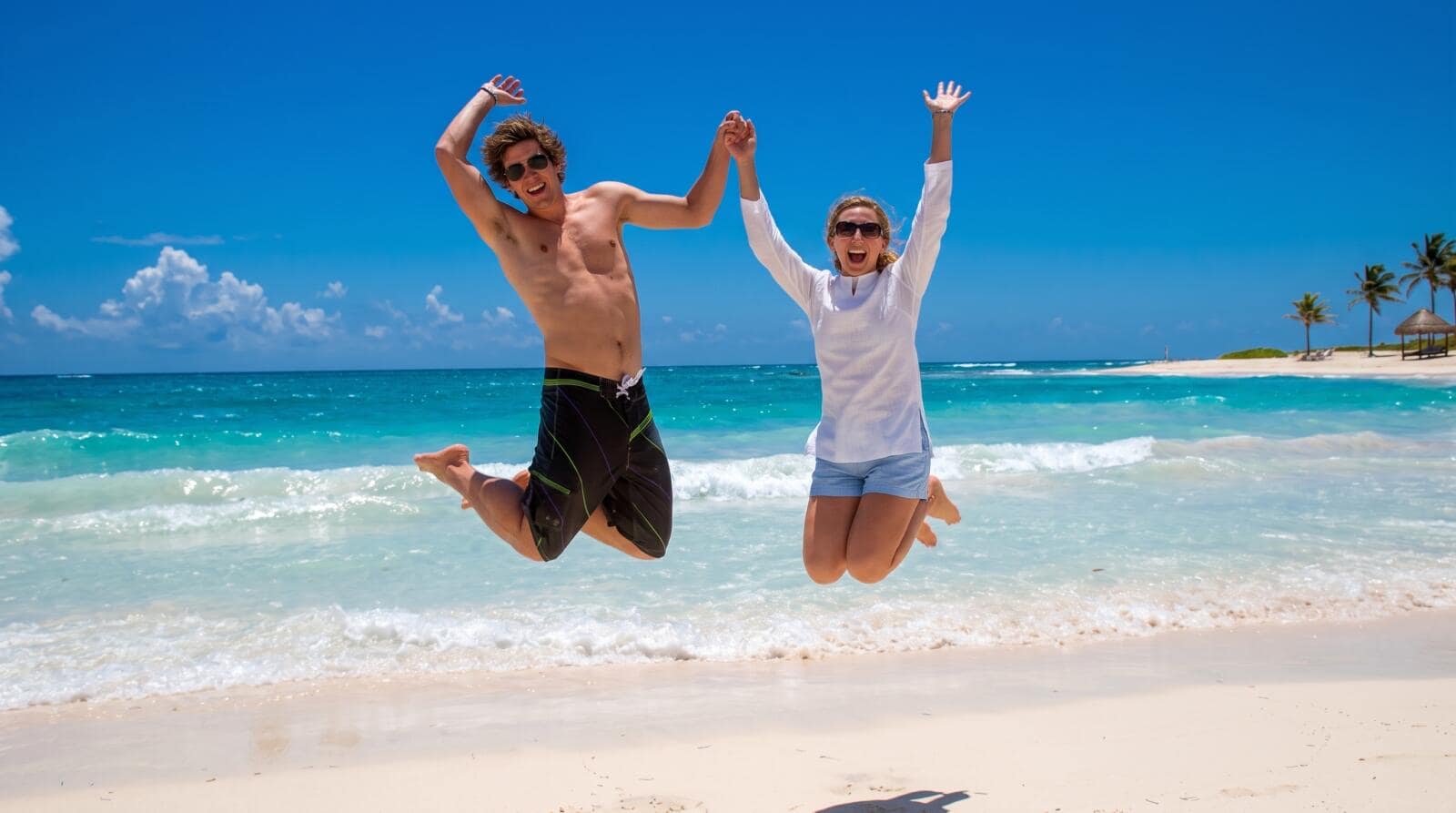 A joyful man and woman holding hands while jumping high into the air on a white sand beach, with vibrant turquoise sea water and palm trees behind them under a bright blue sky.