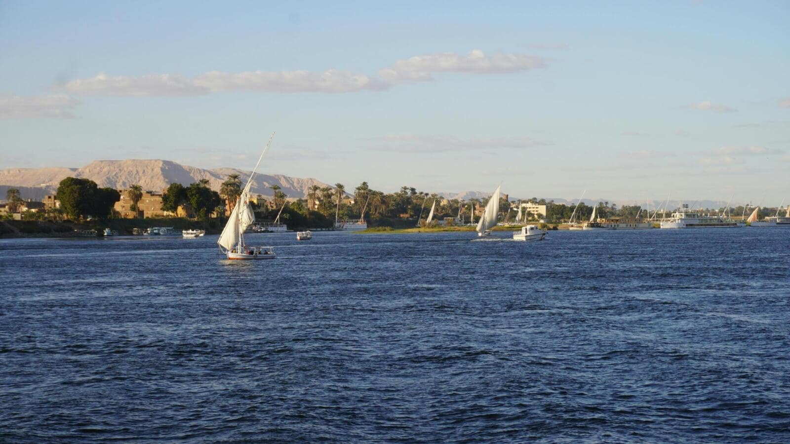 Traditional Egyptian felucca sailboats sailing on the Nile River in Luxor with lush riverbanks and desert hills in the background.9 Days Cairo and Nile cruise.