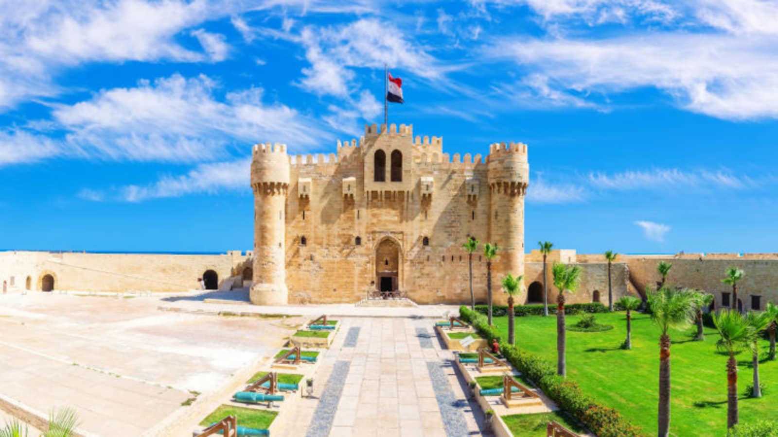 A wide daytime view of the prominent stone fortress of the Citadel of Qaitbay in Alexandria, featuring its fortified towers, main entrance, and a landscaped green garden under a bright blue sky with light clouds.