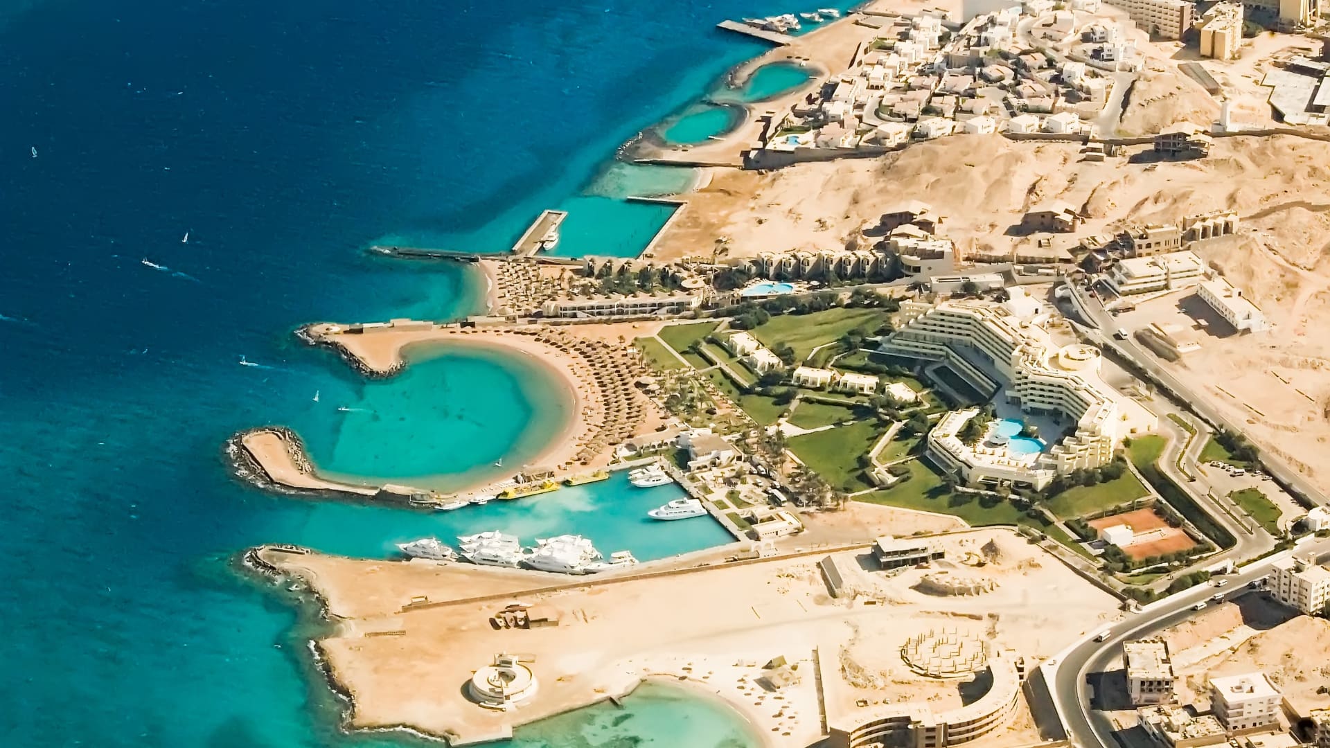 A high-angle aerial view of the Red Sea coastline in Hurghada, showcasing deep blue waters shifting to a turquoise lagoon beach and a premium resort hotel layout along the coast.