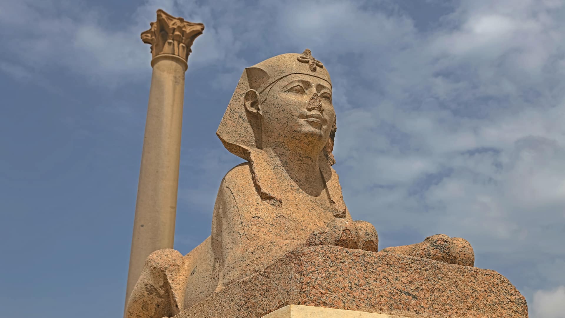 The ancient Roman Pompey's Pillar alongside a granite Sphinx statue under a blue sky in Alexandria Egypt.