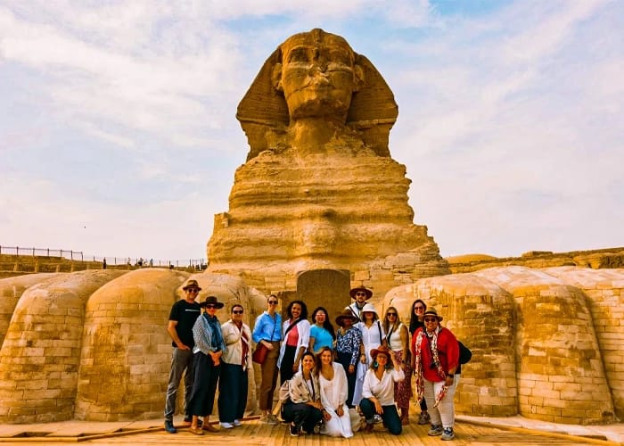 A diverse group of travelers smiling and posing together directly in front of the monumental Great Sphinx on the Giza Plateau under a soft daytime sky.