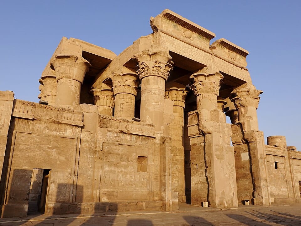 A warm, low-angle daytime view of the ancient stone portal and heavy carved columns of the Temple of Kom Ombo against a clear sky.